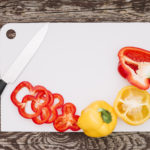 Wooden worktop with white melamine kitchen boards on. Slices of red and yellow peppers and a sharp knife are placed on top.