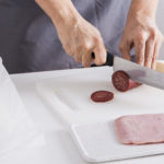 Man preparing salami on white melamine kitchen boards with a sharp knife.