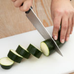 Close up of a woman's hands using a sharp knife to slice a courgette on a white kitchen board.